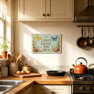 May include: A rectangular metal sign with the text "FAIRIES ENCHANT THIS AREA" and illustrations of a yellow and blue butterfly, surrounded by red and pink flowers. The sign is mounted on a light-colored wall in a kitchen setting.