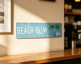 May include: A rectangular light blue metal sign with a distressed finish featuring the white text BEACH BUM and BLVD. The sign includes a graphic of two green palm trees with brown trunks positioned between the words on the right side.