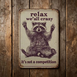 May include: A rectangular metal sign featuring a raccoon illustration. The sign has the text "relax we'all crazy it's not a competition" in a vintage style. The raccoon is depicted with its arms raised.