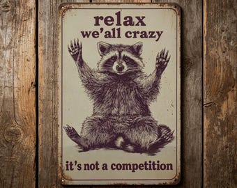May include: A rectangular metal sign with a raccoon illustration. The raccoon is sitting with its arms raised, and the sign has the text "relax we'all crazy it's not a competition" in a vintage style.