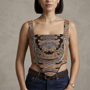 Puede incluir: Top corto de cuello cuadrado con estampado de cachemira en tonos naranja, azul y negro. Se combina con vaqueros oscuros y un cinturón marrón. Un reloj dorado y un delicado collar completan el look.