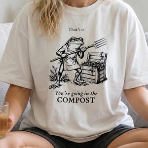 Op de afbeelding: Crèmekleurig T-shirt met een zwart-witte illustratie van een kikker met een hooivork, met de tekst "That's it." erboven en "You're going in the COMPOST" eronder. Het shirt is een casual, comfortabele stijl.
