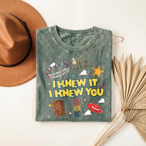 Op de afbeelding: Een mineraal gewassen saliegroen t-shirt met de tekst "I KNEW IT I KNEW YOU" in het geel, samen met afbeeldingen van een kralenarmband, een sheriffster, cowboylaarzen, letterblokken en een rode hoed, met de tekst "infinity and beyond".