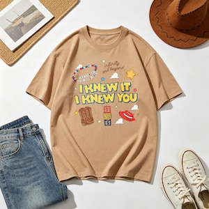 Op de afbeelding: Een beige t-shirt met korte mouwen en de tekst I KNEW IT I KNEW YOU in het geel met blauwe omlijning. De afbeelding bevat een kralenarmband, een gele ster, een bruine cowboylaars, een stapel blokken met de letters T, S, 5 en een rode hoed.