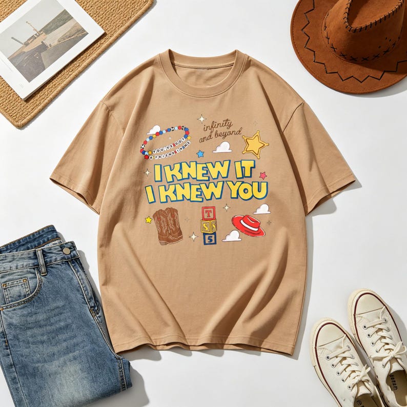 Op de afbeelding: Een beige t-shirt met korte mouwen en de tekst I KNEW IT I KNEW YOU in het geel met blauwe omlijning. De afbeelding bevat een kralenarmband, een gele ster, een bruine cowboylaars, een stapel blokken met de letters T, S, 5 en een rode hoed.