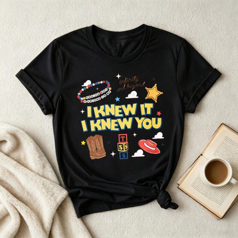 Op de afbeelding: Een zwart t-shirt met korte mouwen en een grafische print met de tekst I KNEW IT I KNEW YOU, infinity and beyond, en YOU'VE GOT A FRIEND IN ME. Het ontwerp bevat illustraties van een kralenarmband, een ster, cowboylaarzen, blokken en een hoed.