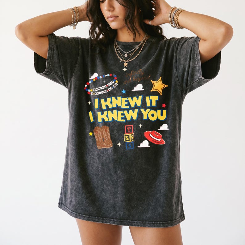 Op de afbeelding: Een antracietgrijs acid-wash t-shirt met de gele tekst I KNEW IT I KNEW YOU. De afbeelding bevat een kleurrijke kralenarmband, een gouden sheriffster, bruine cowboylaarzen, een rode hoed en een stapel blokken met de letters T S S.