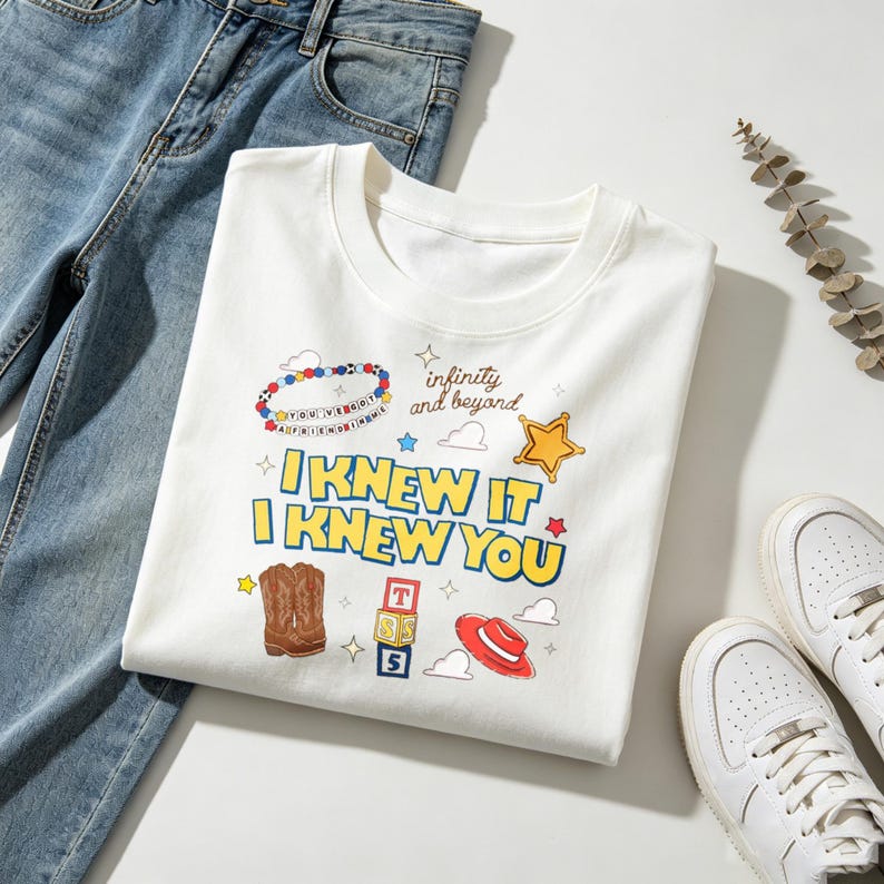 Op de afbeelding: Een wit t-shirt met een grafische print met de tekst "I KNEW IT I KNEW YOU", "infinity and beyond" en "YOU'VE GOT A FRIEND IN ME" op een kralenarmband, naast illustraties van cowboylaarzen, een ster, blokken en een hoed.