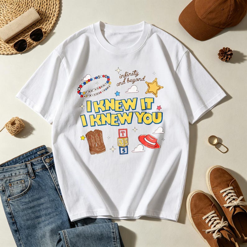 Op de afbeelding: Een wit t-shirt met korte mouwen en de tekst I KNEW IT I KNEW YOU in het geel met blauwe omlijning. De afbeelding bevat een kralenarmband, een gele ster, bruine cowboylaarzen, alfabetblokken en een rode hoed met de tekst infinity and beyond.