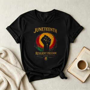 Op de afbeelding: Een zwart t-shirt met korte mouwen en een afbeelding van een opgeheven vuist in het midden van een rode, gele en groene zonnestraal. De tekst luidt JUNETEENTH, JUNE 19, 1865, RESILIENT FREEDOM, UNBROKEN, UNSHAKEN, UNSTOPPABLE, met een klein leeuwembleem.