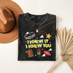 Op de afbeelding: Een antracietgrijs, mineraal gewassen t-shirt met de gele tekst I KNEW IT I KNEW YOU, een kralenarmband-afbeelding met de tekst YOU'VE GOT A FRIEND IN ME, een cowboylaars, een rode hoed, alfabetblokken, een gouden ster en kleine wolkjes.