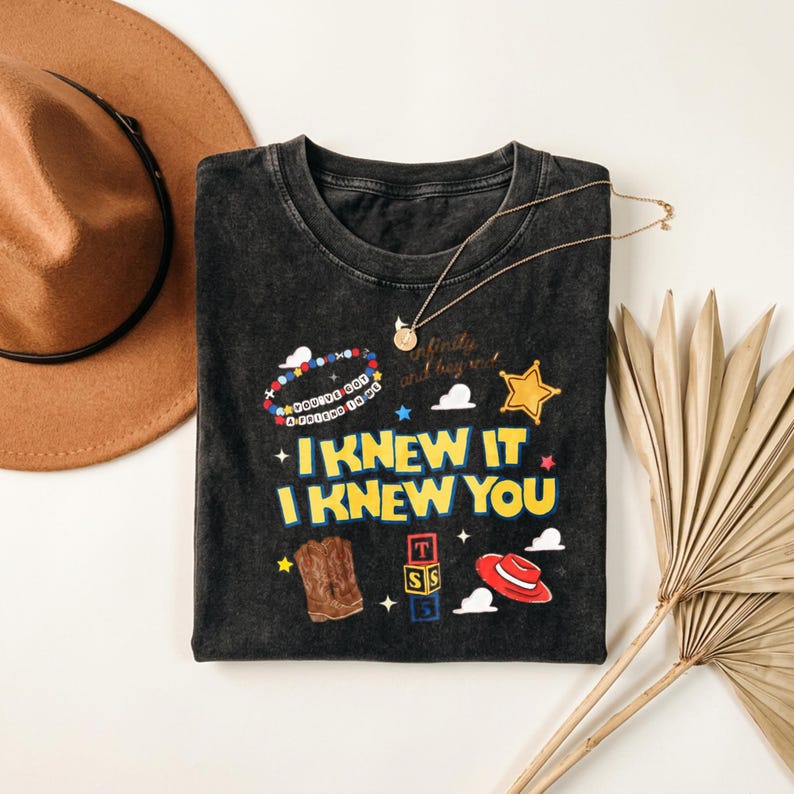 Op de afbeelding: Een antracietgrijs, mineraal gewassen t-shirt met de gele tekst I KNEW IT I KNEW YOU, een kralenarmband-afbeelding met de tekst YOU'VE GOT A FRIEND IN ME, een cowboylaars, een rode hoed, alfabetblokken, een gouden ster en kleine wolkjes.