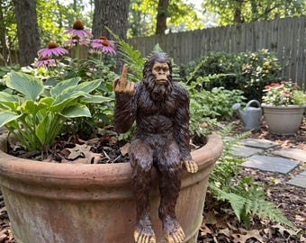 May include: A brown, textured Bigfoot figurine with fur detailing, sitting on the rim of a large terracotta planter. The figurine is depicted with a serious facial expression and is holding up its middle finger in a distinct gesture.