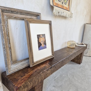 Puede incluir: Un banco rústico de madera marrón oscuro con una parte superior rectangular y una base de bloque. Dos cuadros enmarcados se apoyan en la parte trasera del banco. Una pequeña alfombra decorativa cuelga de la pared detrás del banco.