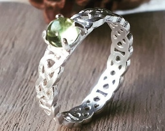 Può includere: Un anello d'argento con una gemma verde. L'anello presenta un design traforato dettagliato lungo la fascia. La gemma rotonda è incastonata in una montatura a griffe. L'anello è esposto su una superficie di legno.