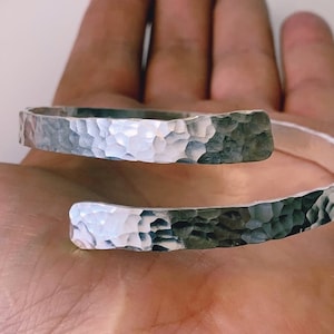 May include: A silver hammered bangle bracelet with a simple, open design. The bracelet is made from a single piece of metal that is bent into a circle and then hammered to create a textured surface.
