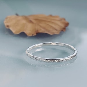 May include: A silver ring with a delicate, etched pattern. The ring has a thin band and a slightly hammered texture. The design features a repeating, swirling motif. A dried leaf is in the background.