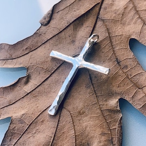 May include: A silver hammered cross pendant on a dried brown leaf.