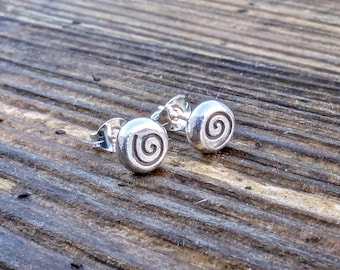 May include: A pair of small, round, silver-colored stud earrings, each featuring a centered, engraved spiral design. The earrings have a polished, metallic finish and include standard post-style backings for secure attachment to the earlobe.