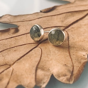 May include: A pair of silver stud earrings with green moss agate stones. The earrings are on a brown leaf.