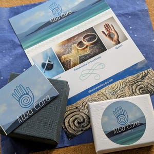 May include: Two small boxes with the Alba Cara Jewellery logo. The boxes are on a blue background with a printed card featuring the Alba Cara Jewellery logo, a cross, and the text "Handmade in Scotland".