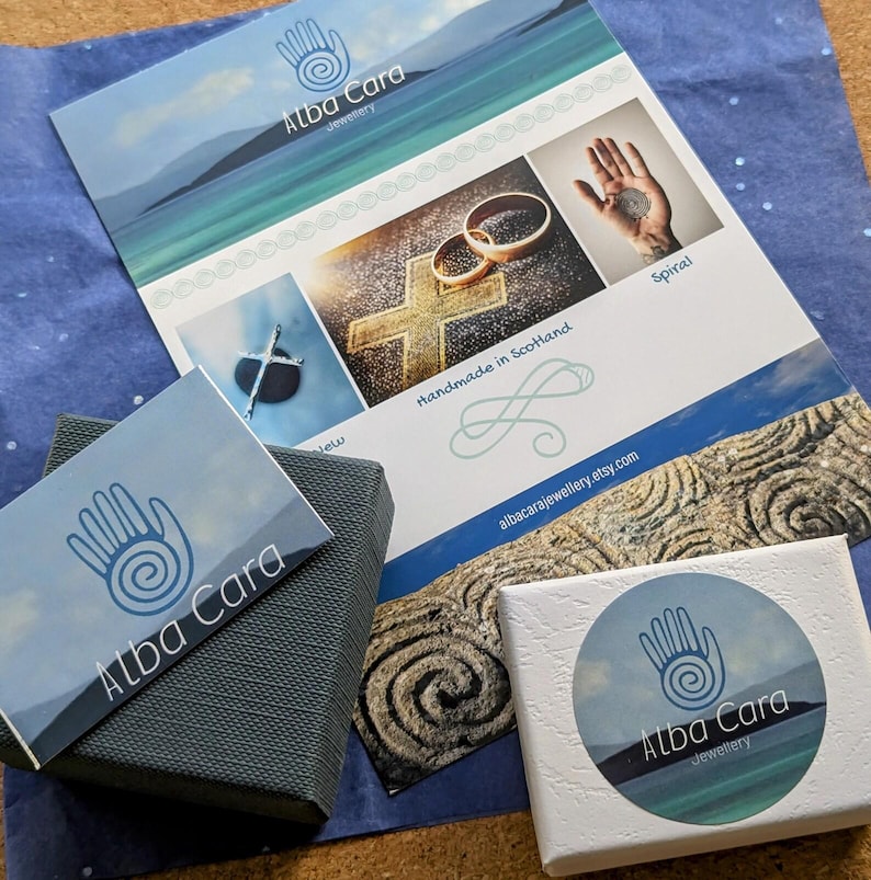 May include: Two small boxes with the Alba Cara Jewellery logo. The boxes are on a blue background with a printed card featuring the Alba Cara Jewellery logo, a cross, and the text "Handmade in Scotland".