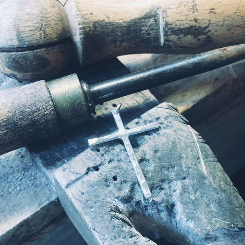 May include: A silver cross pendant with a textured surface, resting on a wooden workbench. The cross is a popular religious symbol and is often used in jewelry.