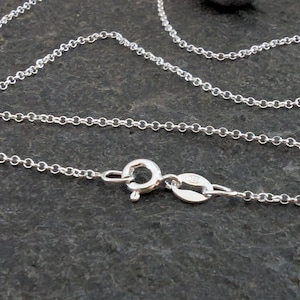 May include: Close-up of a delicate silver chain necklace with a lobster clasp. The chain is made of small, interconnected circular links. The clasp is open, ready to be fastened. The necklace rests on a dark, textured surface.