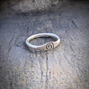 May include: A polished silver ring featuring a smooth, rounded band that tapers into a flat, circular face. The centre of the face is engraved with a small, dark, spiralling pattern that contrasts against the bright, reflective metallic surface.