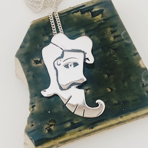 May include: A silver pendant necklace featuring a stylized profile of a person with a beard and wavy hair. The pendant hangs from a delicate silver chain. The pendant is displayed on a dark green, textured surface.