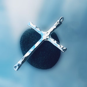 May include: A silver hammered cross pendant resting on a dark gray stone. The cross has a textured surface and a simple design.