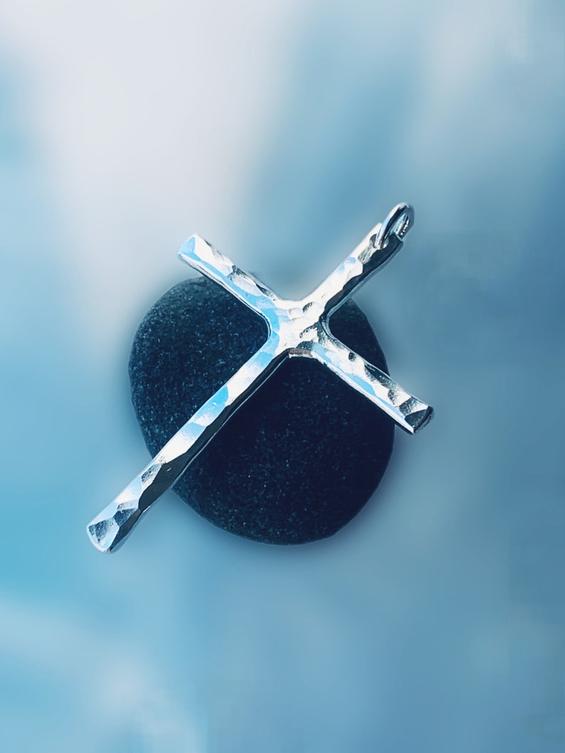 May include: A silver hammered cross pendant resting on a dark gray stone. The cross has a textured surface and a simple design.
