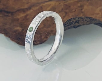 Può includere: Un anello d'argento con un motivo floreale inciso e una piccola gemma verde. L'anello è posto su uno sfondo di legno naturale, evidenziando il suo design e la sua maestria. Un gioiello.