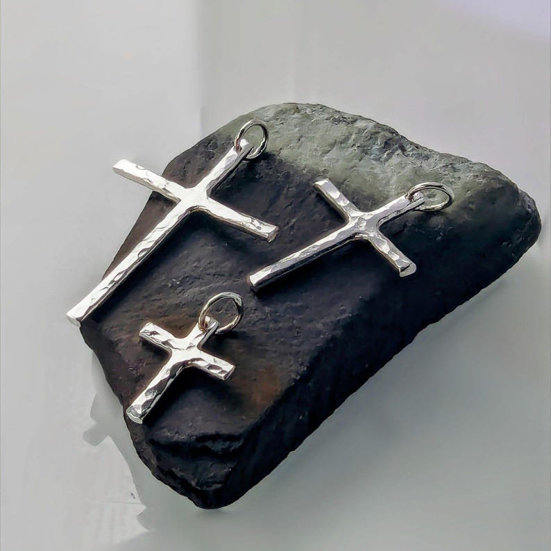 May include: Three silver cross pendants of varying sizes, displayed on a dark gray rock. The crosses have a hammered texture.