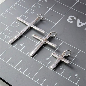 May include: Three silver cross pendants of different sizes, laid out on a gray grid with white lines and numbers. The crosses are hammered and have a textured surface.