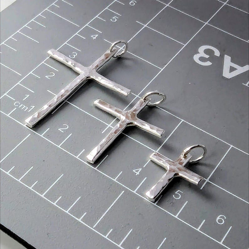 May include: Three silver cross pendants of different sizes, laid out on a gray grid with white lines and numbers. The crosses are hammered and have a textured surface.