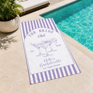 May include: A white and lavender striped beach towel with the text "THE BRIDE Club" and "Doki's Bachelorette Las Vegas, Nevada". The towel features a graphic of two martini glasses.