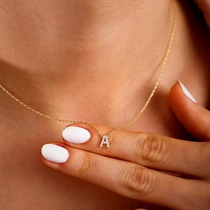 May include: A delicate gold necklace featuring a small, sparkling letter "A" pendant. The pendant is encrusted with small, clear gemstones. The necklace is worn around the neck, and the chain is visible.