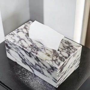 Puede incluir: Un dispensador de pañuelos rectangular de mármol con un diseño veteado blanco y morado oscuro. Los pañuelos blancos sobresalen parcialmente de la abertura superior. La caja está sobre una superficie negra.
