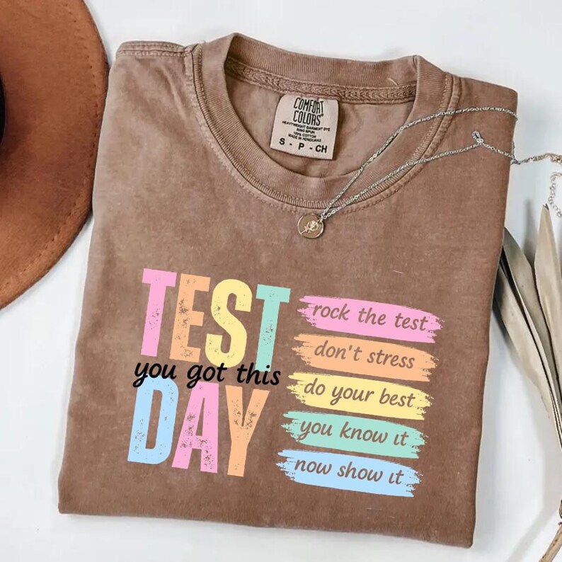 May include: Brown t-shirt with the words "TEST DAY" in colorful block letters. The shirt also has the phrases "rock the test," "don't stress," "do your best," "you know it," and "now show it." A silver necklace with a pendant is on the shirt.