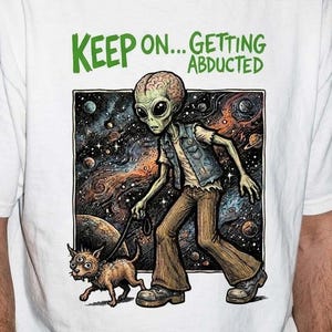 Könnte beinhalten: Weißes T-Shirt mit einer Grafik eines Aliens, der einen Hund im Weltraum ausführt, mit dem grünen Text "KEEP ON... GETTING ABDUCTED". Der Außerirdische trägt eine Weste und eine braune Hose. Der Hund ist klein und braun.