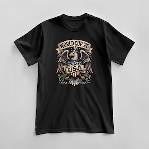 May include: Black t-shirt featuring a graphic of an eagle wearing a hat with the American flag design. The eagle has outstretched wings and holds a shield with the text "Team USA". The text "World Cup '26" is above the eagle.