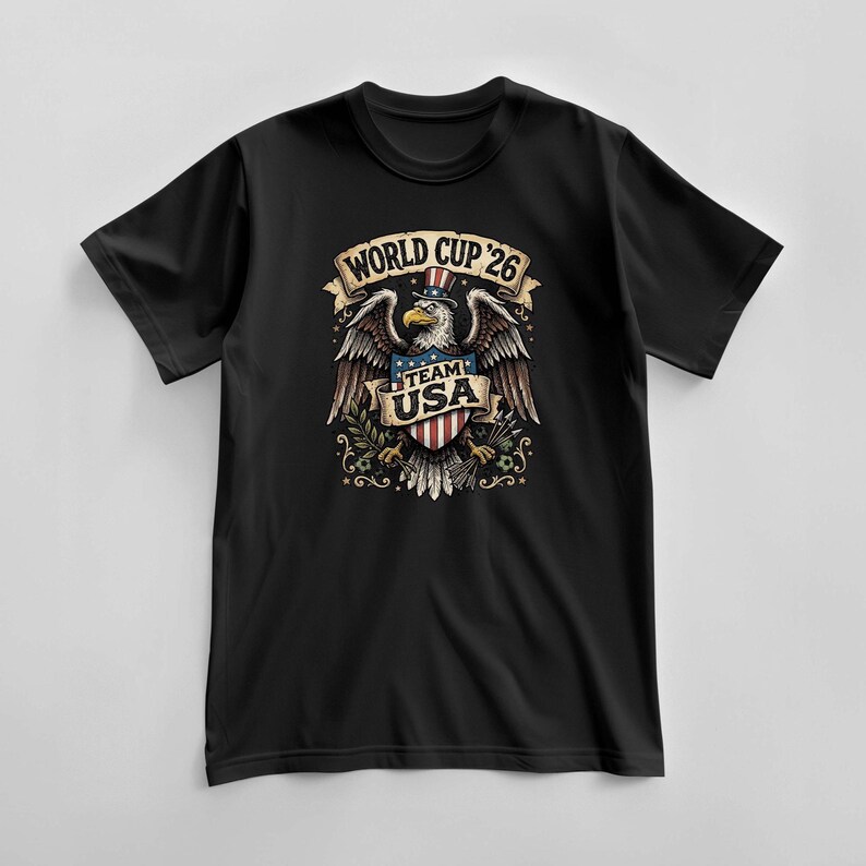 May include: Black t-shirt featuring a graphic of an eagle wearing a hat with the American flag design. The eagle has outstretched wings and holds a shield with the text "Team USA". The text "World Cup '26" is above the eagle.