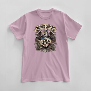 May include: A light pink t-shirt featuring a graphic of an eagle wearing a top hat with the text "WORLD CUP '26" above and "TEAM USA" below. The eagle is holding a shield with red and white stripes.