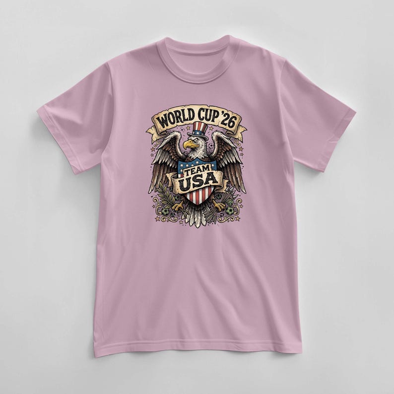 May include: A light pink t-shirt featuring a graphic of an eagle wearing a top hat with the text "WORLD CUP '26" above and "TEAM USA" below. The eagle is holding a shield with red and white stripes.