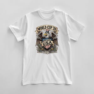 May include: White t-shirt featuring a detailed graphic of an eagle wearing a top hat with the text "WORLD CUP '26" and "TEAM USA". The eagle is holding a shield with the American flag design.
