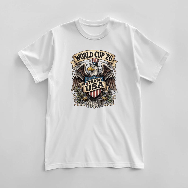 May include: White t-shirt featuring a detailed graphic of an eagle wearing a top hat with the text "WORLD CUP '26" and "TEAM USA". The eagle is holding a shield with the American flag design.