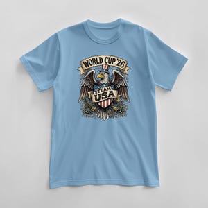 May include: Light blue t-shirt featuring a graphic of an eagle wearing a top hat, with the text "WORLD CUP '26" above and "TEAM USA" below. The eagle is holding a shield with red and white stripes. The shirt is a short-sleeved crew neck.