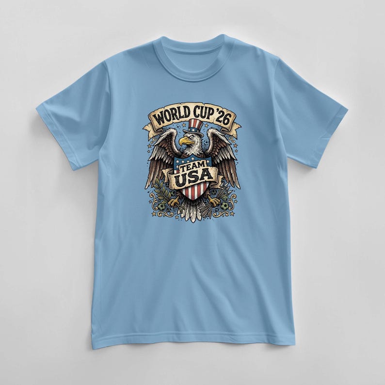 May include: Light blue t-shirt featuring a graphic of an eagle wearing a top hat, with the text "WORLD CUP '26" above and "TEAM USA" below. The eagle is holding a shield with red and white stripes. The shirt is a short-sleeved crew neck.