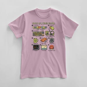 May include: A light pink t-shirt featuring a graphic of video editing tools. The design includes a timeline, keyboard, mouse, headphones, coffee mug, hard drive, monitor, meter, and an "export" button. The text "TOOLS OF A VIDEO EDITOR" is at the top.